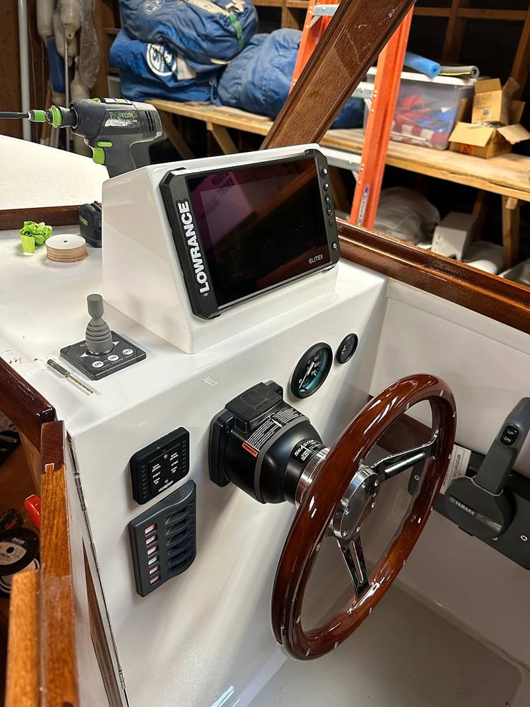 A white helm console with a chartplotter pod, joystick, switch panels and a wooden wheel